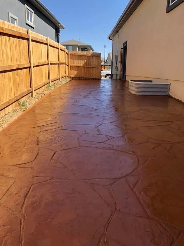 Stamped concrete pathway alongside a wooden fence and house under a clear blue sky.