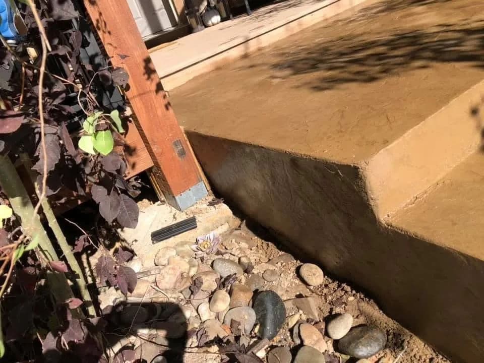 Concrete patio with sloped edge, surrounded by pebble landscaping and plants.