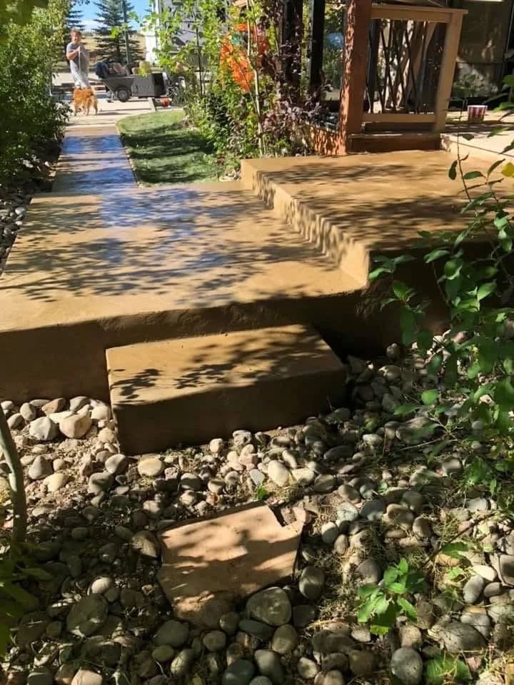 Backyard pathway with steps, surrounded by stones and greenery, sunlight casting shadows.