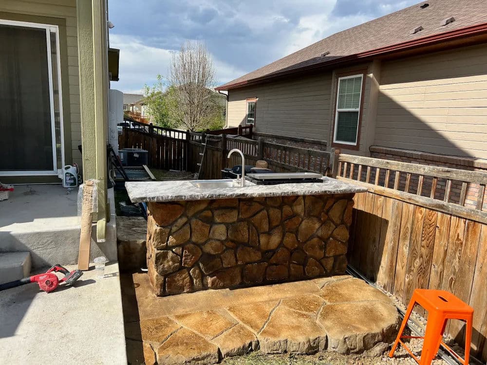 Outdoor stone countertop with sink in backyard, surrounded by wooden fence and houses.