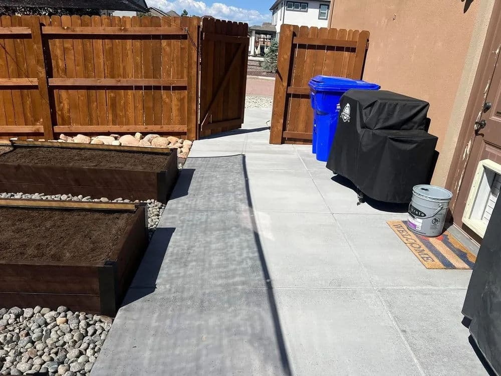 Image of a backyard with garden beds, stone path, blue trash bins, and a grill near a wooden fence.
