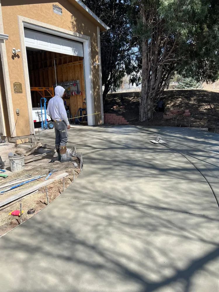 Worker finishing a newly poured concrete driveway near a garage under tree shade.