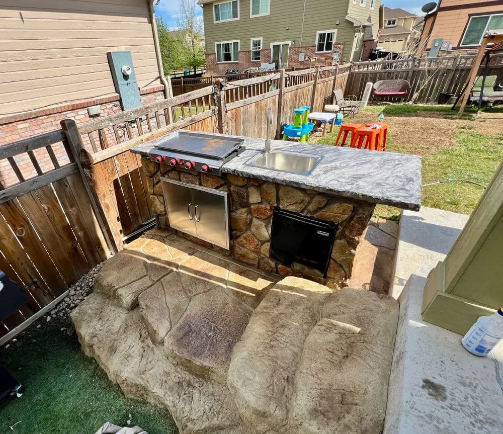 Outdoor kitchen featuring a stone grill station with a sink and refrigerator in a backyard.