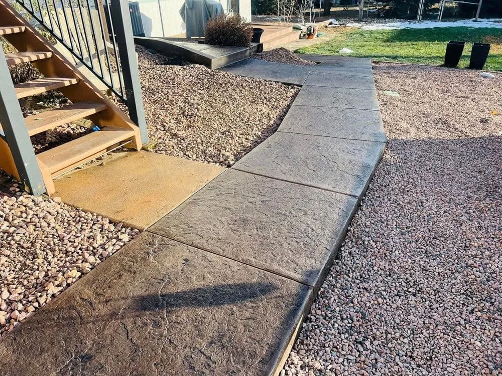 Textured concrete walkway leading to stairs in a landscaped yard with gravel and grass.