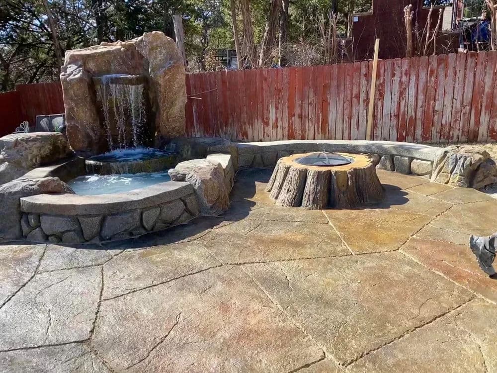 Backyard oasis featuring a stone waterfall, hot tub, and rustic tree stump seating area.