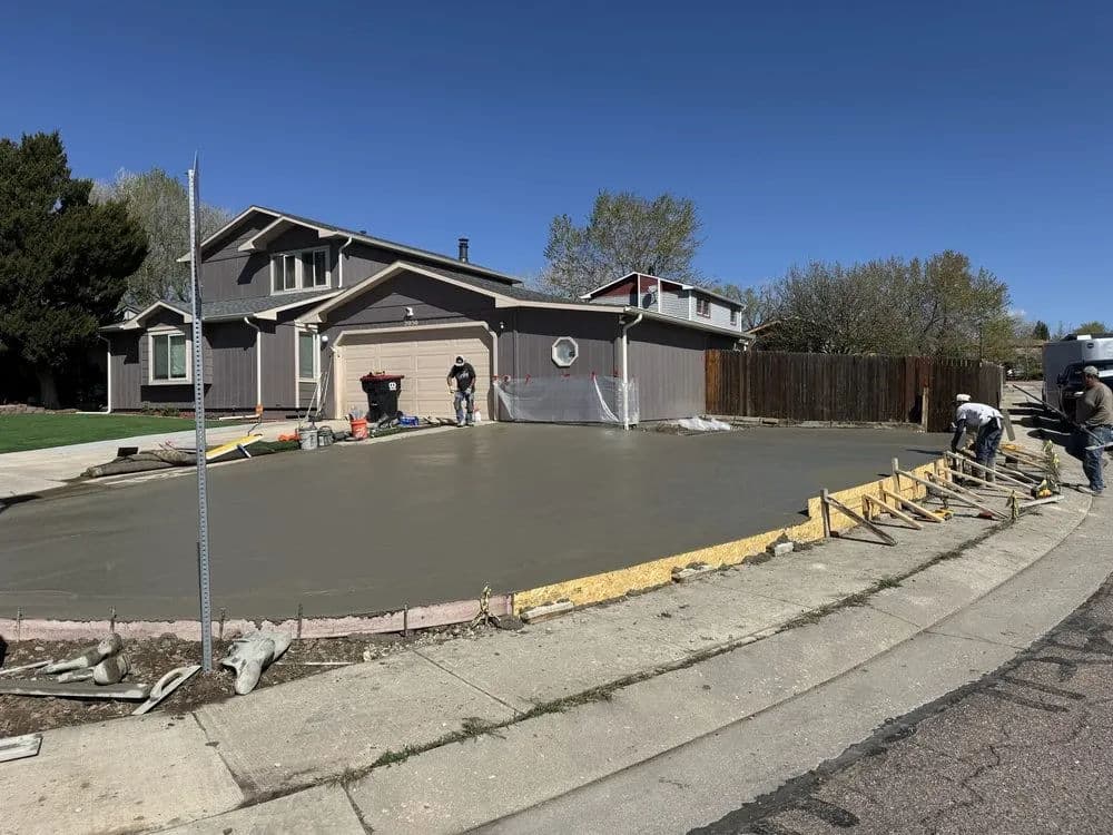 Workers finishing a freshly poured concrete driveway near a suburban home on a clear day.